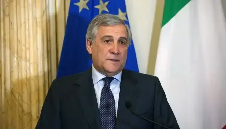 Man in a dark suit speaking at a podium with Italian and European flags.