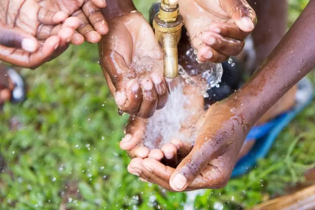 Multiple dark-skinned hands wash under a brass faucet with flowing water outdoors.