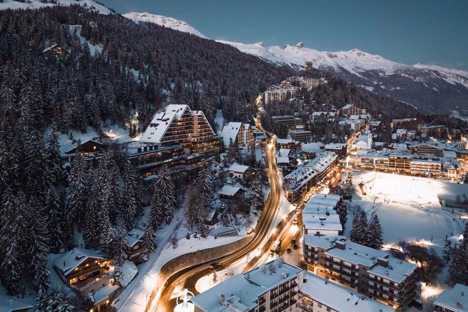 Aerial view of a snowy mountain resort town at night with illuminated buildings and roads.