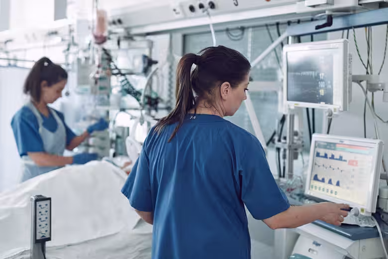 Two nurses caring for a patient in an intensive care unit surrounded by medical monitors.