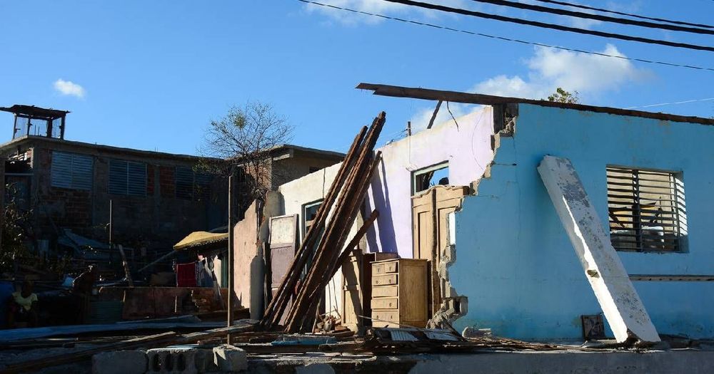 Severely damaged light blue building with collapsed walls and debris after a storm.
