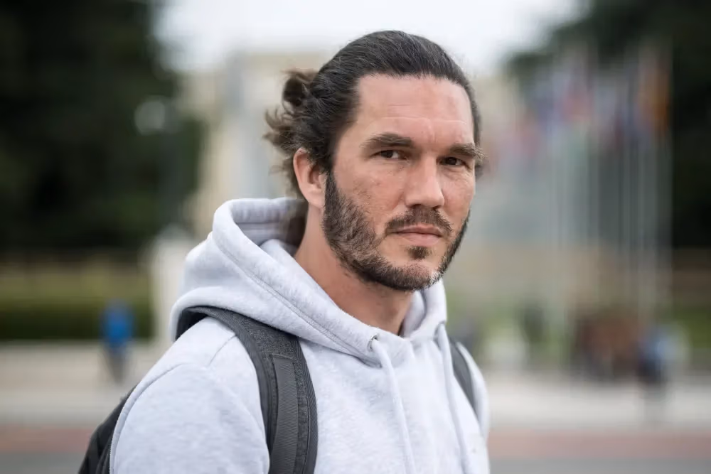 Serious man with dark hair, beard, and grey hoodie looks directly ahead.