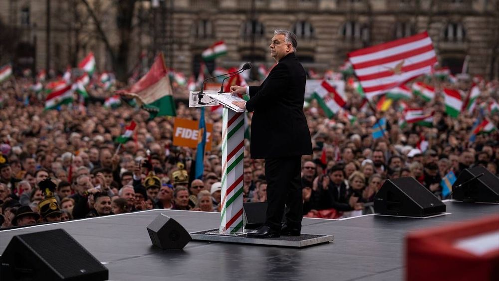 Man speaks from podium to large crowd waving Hungarian flags.
