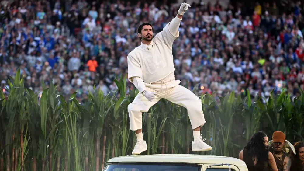 Man in white jumpsuit jumps on vehicle, raising arm during a performance before a crowd.