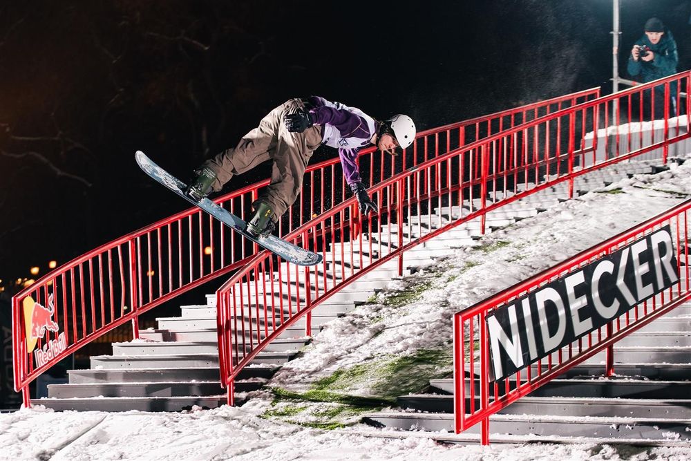 Snowboarder grinding a red Nidecker handrail on a snowy staircase at night.
