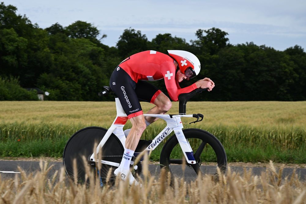 Male cyclist in red suit on white time trial bike in aerodynamic position through a field.
