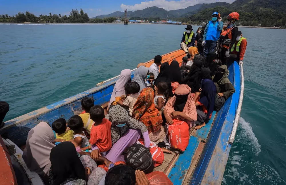 Crowded wooden boat with women and children moves across blue water towards land.