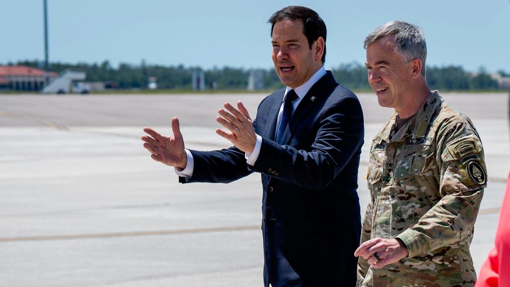Man in suit gestures speaking to military officer on an outdoor airfield.