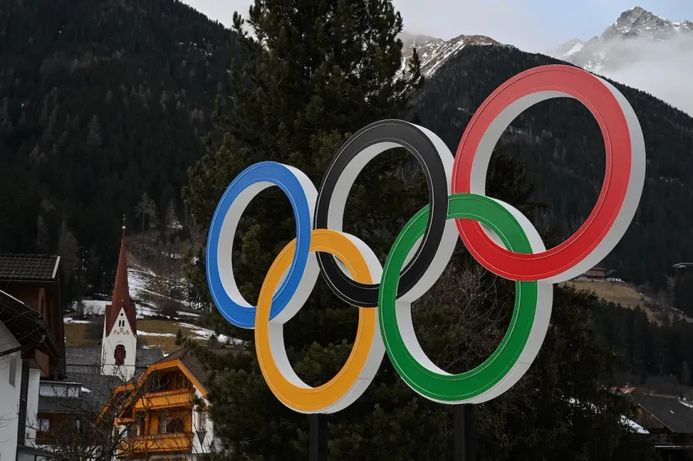 The Olympic rings logo stands prominently against a backdrop of snow-capped mountains and trees.