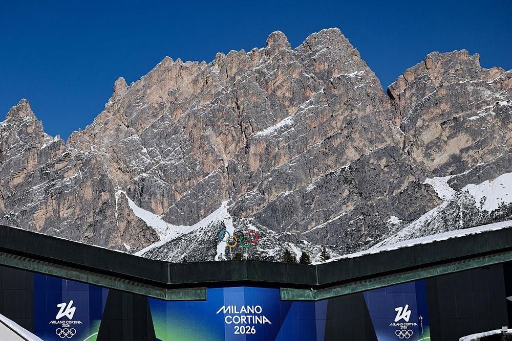 Rugged mountains with snow peaks, clear blue sky, and a "MILANO CORTINA 2026" Olympic banner.