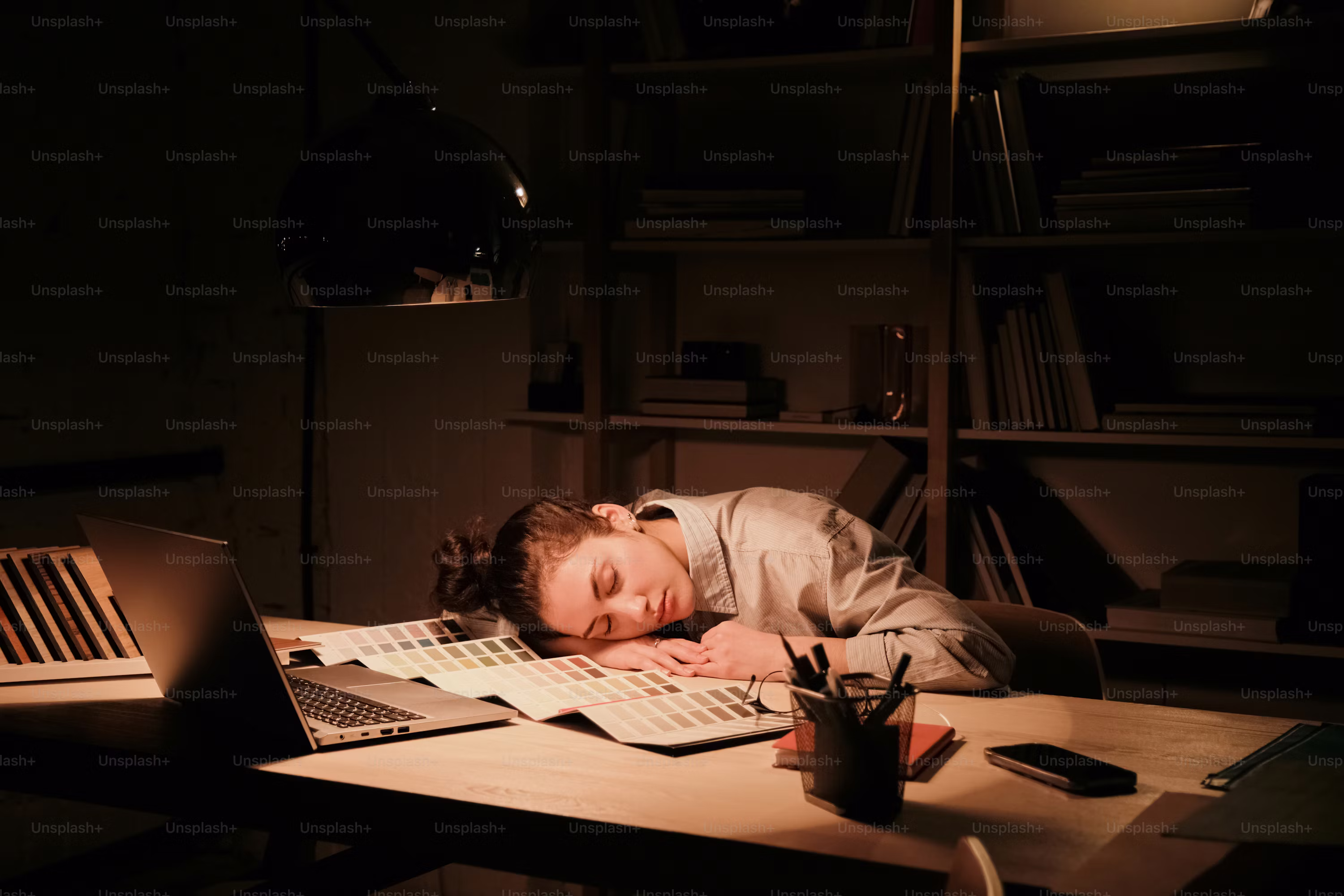 A woman sleeping at a desk, illuminated by a lamp, with color swatches and a laptop.