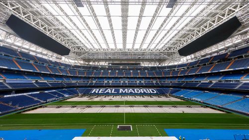 Modern, empty Real Madrid stadium with a retractable roof and blue seating, "REAL MADRID" on field.