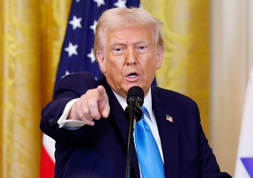 Donald Trump speaking at a podium, pointing to the camera with an American flag behind him.