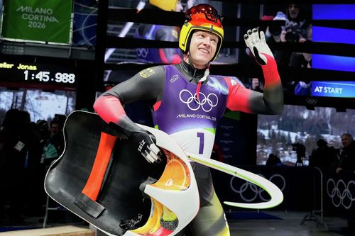 Male luge athlete in a black and red racing suit waves while holding his sled.
