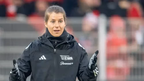 Female soccer coach wearing a black Adidas jacket, gesturing animatedly on the sidelines.