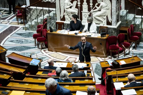 Politician speaking at a podium in a parliamentary chamber with an audience.