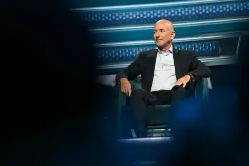 A man in a dark suit sits in a green chair, looking right with a thoughtful expression.