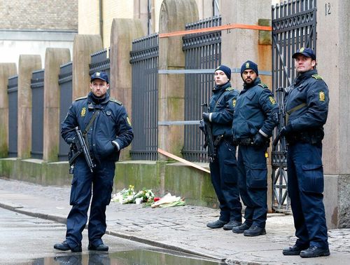Four police officers in dark uniforms stand outside a stone building, one holding a rifle.