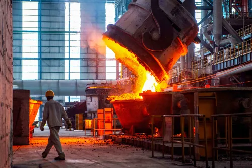 Worker watches molten metal pour from a large ladle in a busy industrial foundry.