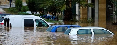 Multiple cars, including a white van, are submerged in deep brown floodwater.