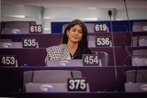 Young woman with patterned scarf smiles from her seat in a legislative assembly.