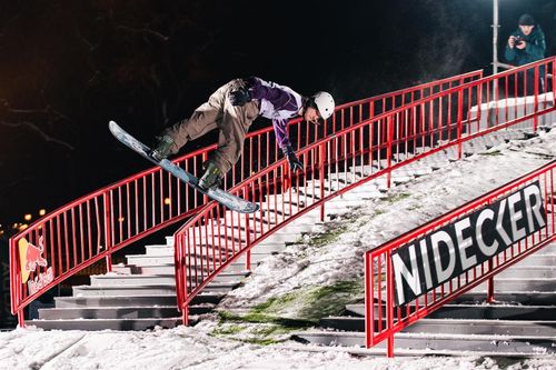 Snowboarder grinding a red Nidecker handrail on a snowy staircase at night.