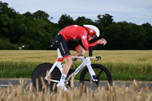 Male cyclist in red suit on white time trial bike in aerodynamic position through a field.
