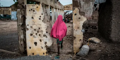 Person in pink hijab walks through bullet-ridden wall in war-torn area.