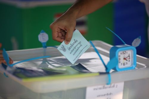 A hand drops a ballot into a transparent ballot box next to a small blue clock.