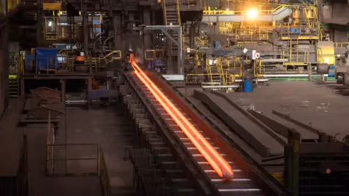 Two glowing hot metal bars move along a conveyor belt in a bustling steel mill.