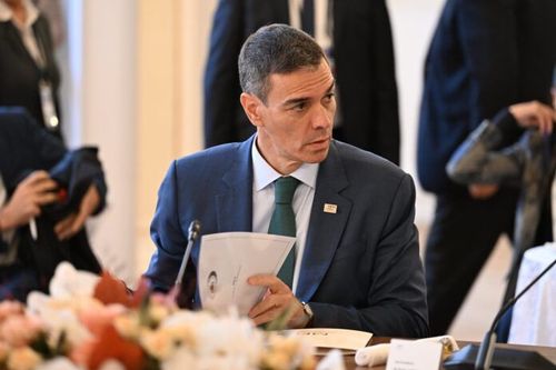 Pedro Sánchez, in a navy suit and green tie, sits at a conference table, looking right.