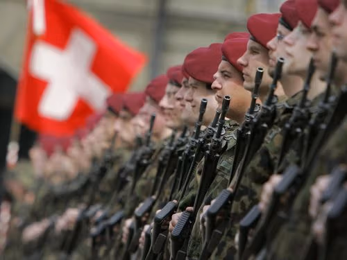 Swiss soldiers in red berets with rifles stand in formation under their national flag.