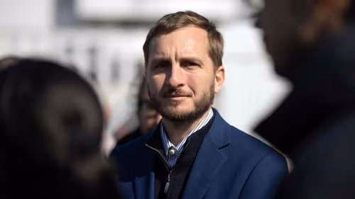 A man with a beard and short brown hair looks intently, wearing a dark blue jacket.