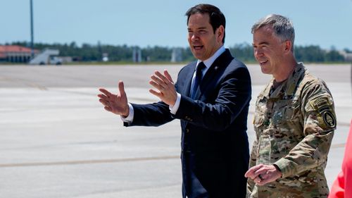 Man in suit gestures speaking to military officer on an outdoor airfield.