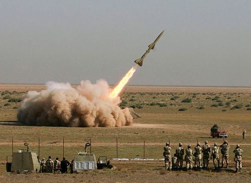 A missile launches from a desert landscape with a large smoke cloud, observed by soldiers.