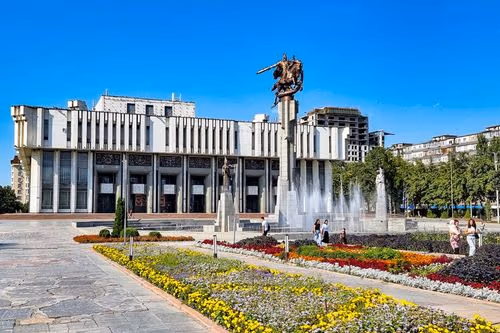 Ornate building with columns, large monument fountain, and vibrant flower garden under blue sky.