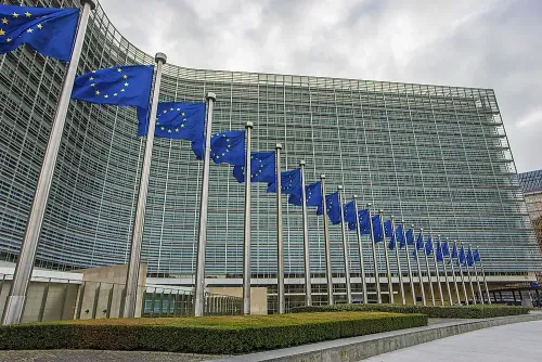 Row of European Union flags in front of a large, modern glass building under an overcast sky.
