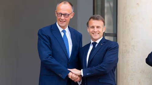 Two smiling men in dark suits shake hands outside a formal building entrance.