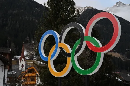 The Olympic rings logo stands prominently against a backdrop of snow-capped mountains and trees.