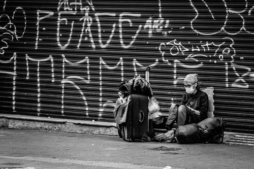 Person in mask sits beside bags against a graffiti-covered wall displaying "PEACE."