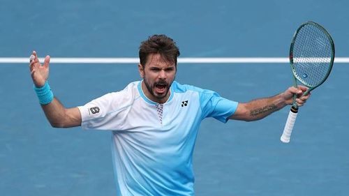 Male tennis player with a beard raises arms on a blue court, racket in hand.