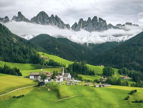 A picturesque village with a church in a green valley beneath jagged, cloud-covered mountains.